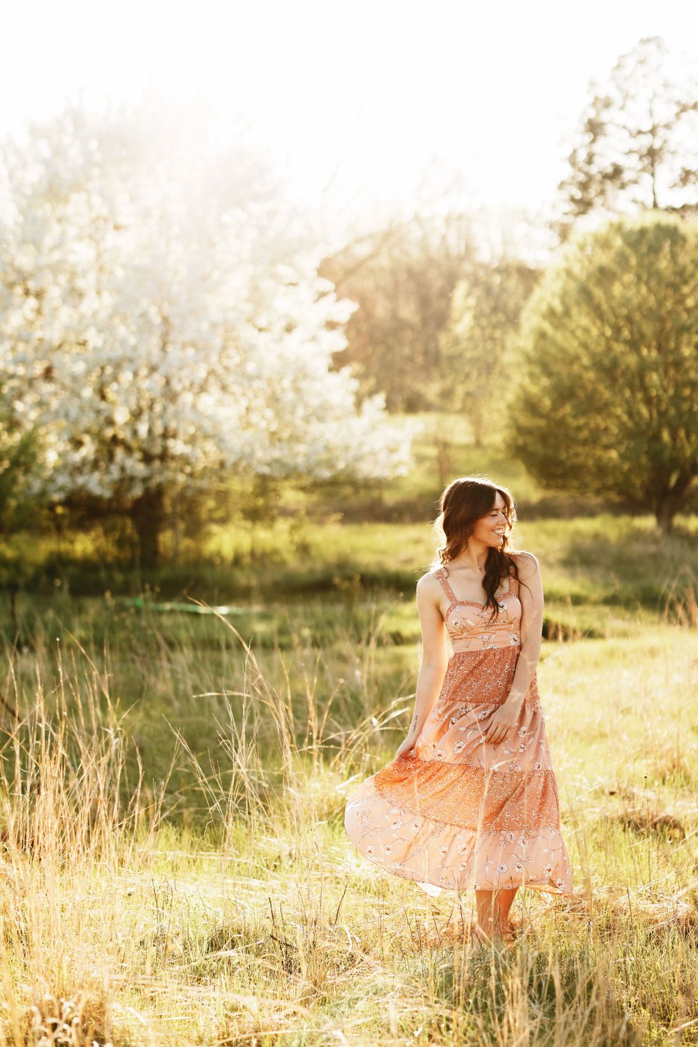 Woman in flowy peach dress twirls in sunlit meadow with wildflowers and trees in background.