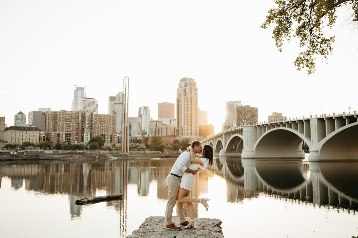 A romantic couple shares a moment at sunset with the Minneapolis skyline and bridge reflecting in the river.