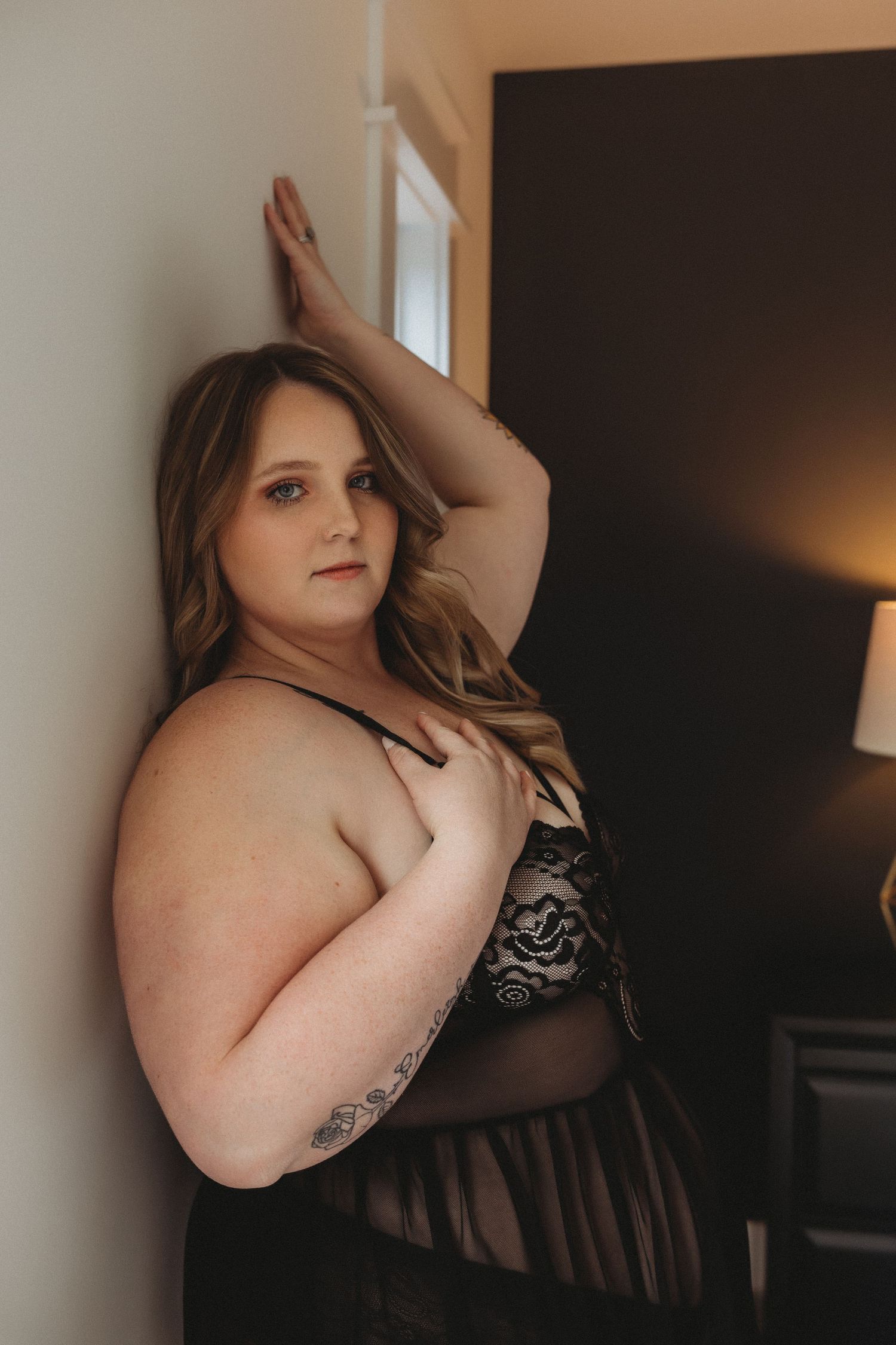 Artistic boudoir photo of person in sheer black dress against light wall.