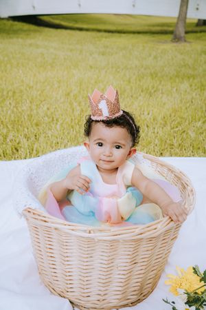 Baby wearing floral crown and pastel dress sits in large wicker basket on grass.