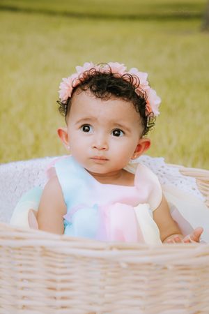 Baby in white and pastel dress peeks over edge of wicker basket outdoors.