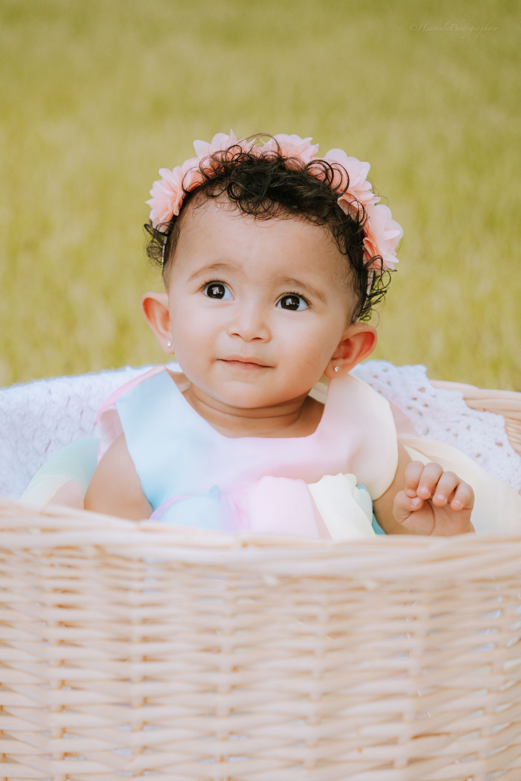 Baby wearing flower crown peers over edge of wicker basket during outdoor photoshoot.