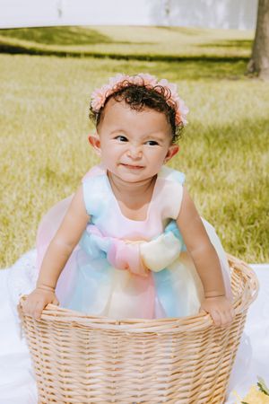 Smiling baby in pastel dress poses inside decorative wicker basket outdoors.