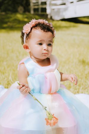 Baby in pastel dress and flower crown plays with flower stem while sitting outdoors.