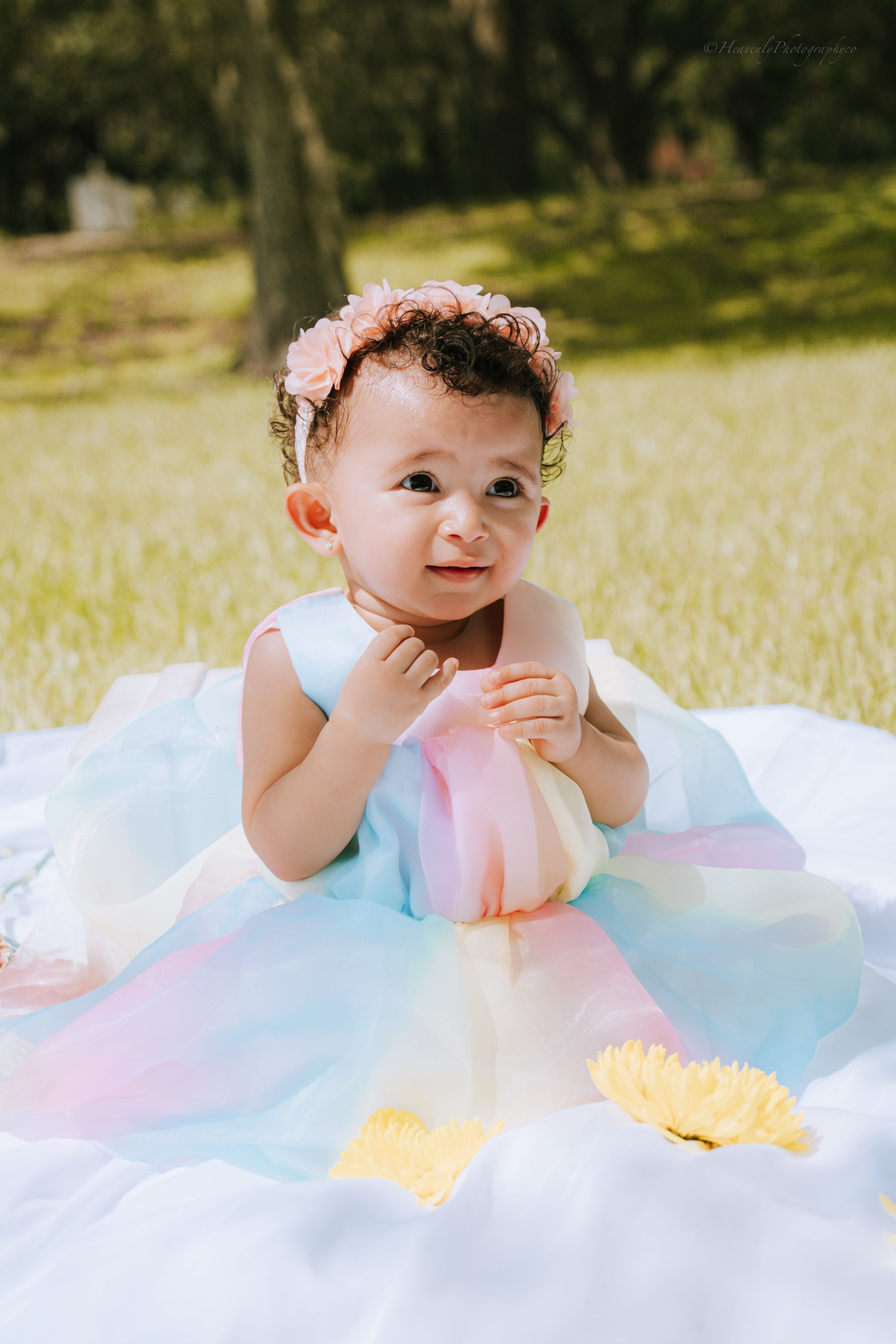 Baby in pastel dress lying on white blanket with floral decorations outdoors.
