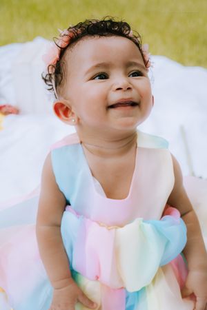 Baby wearing pastel dress with ruffled collar smiles sweetly while lying down outdoors.