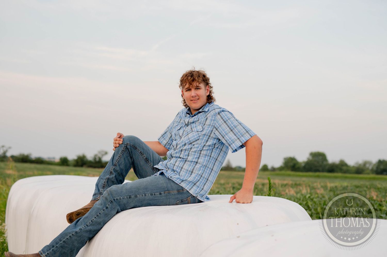 A person in blue plaid lounges on a white wrapped hay bale in a rural field.