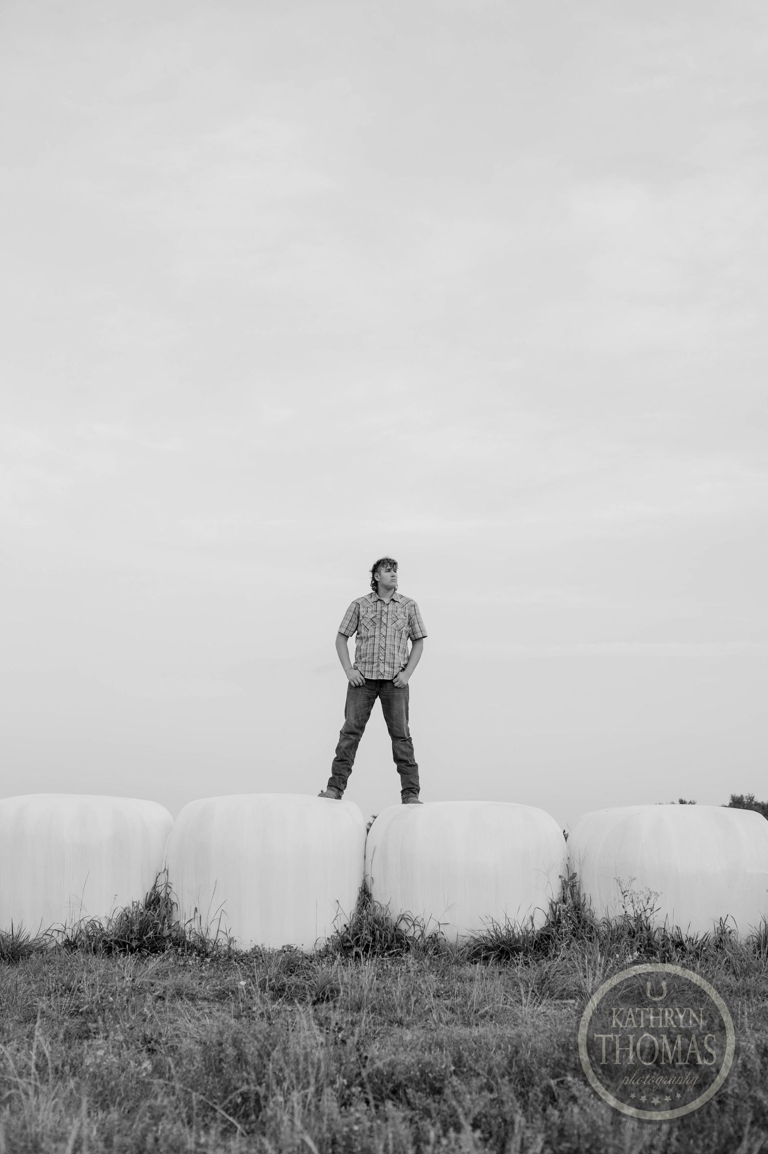 A silhouetted figure stands atop round hay bales in black and white.