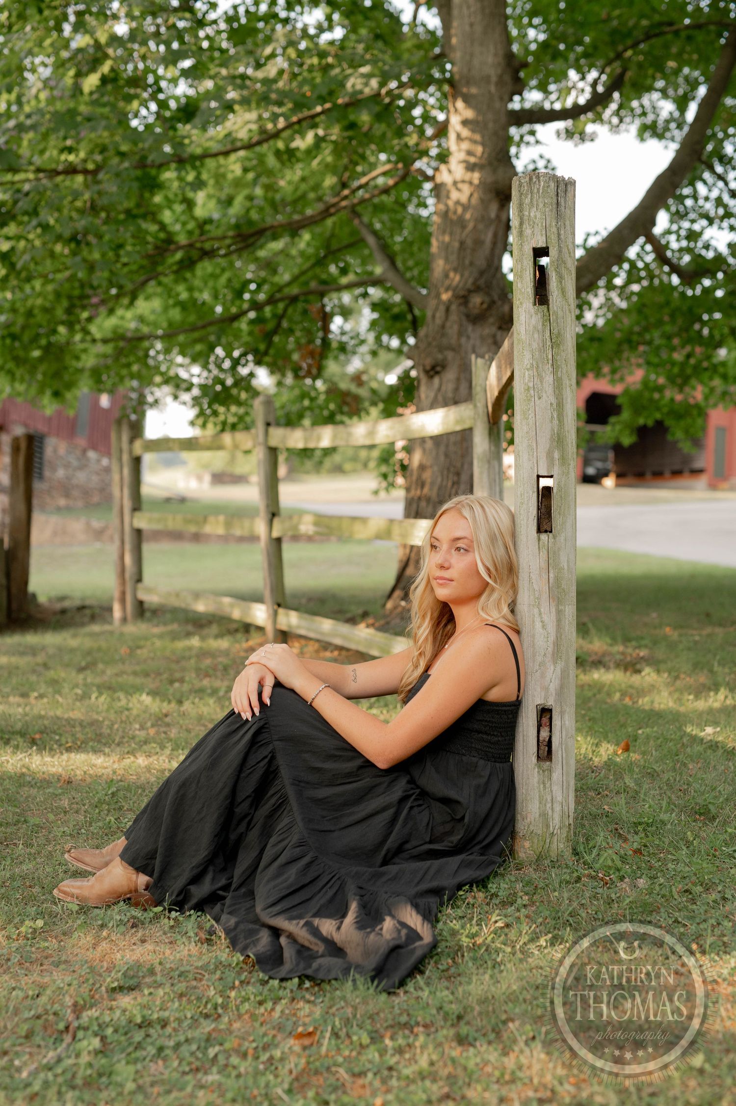 A woman in a flowing black dress sits by a wooden fence post in a rural setting with a barn visible.