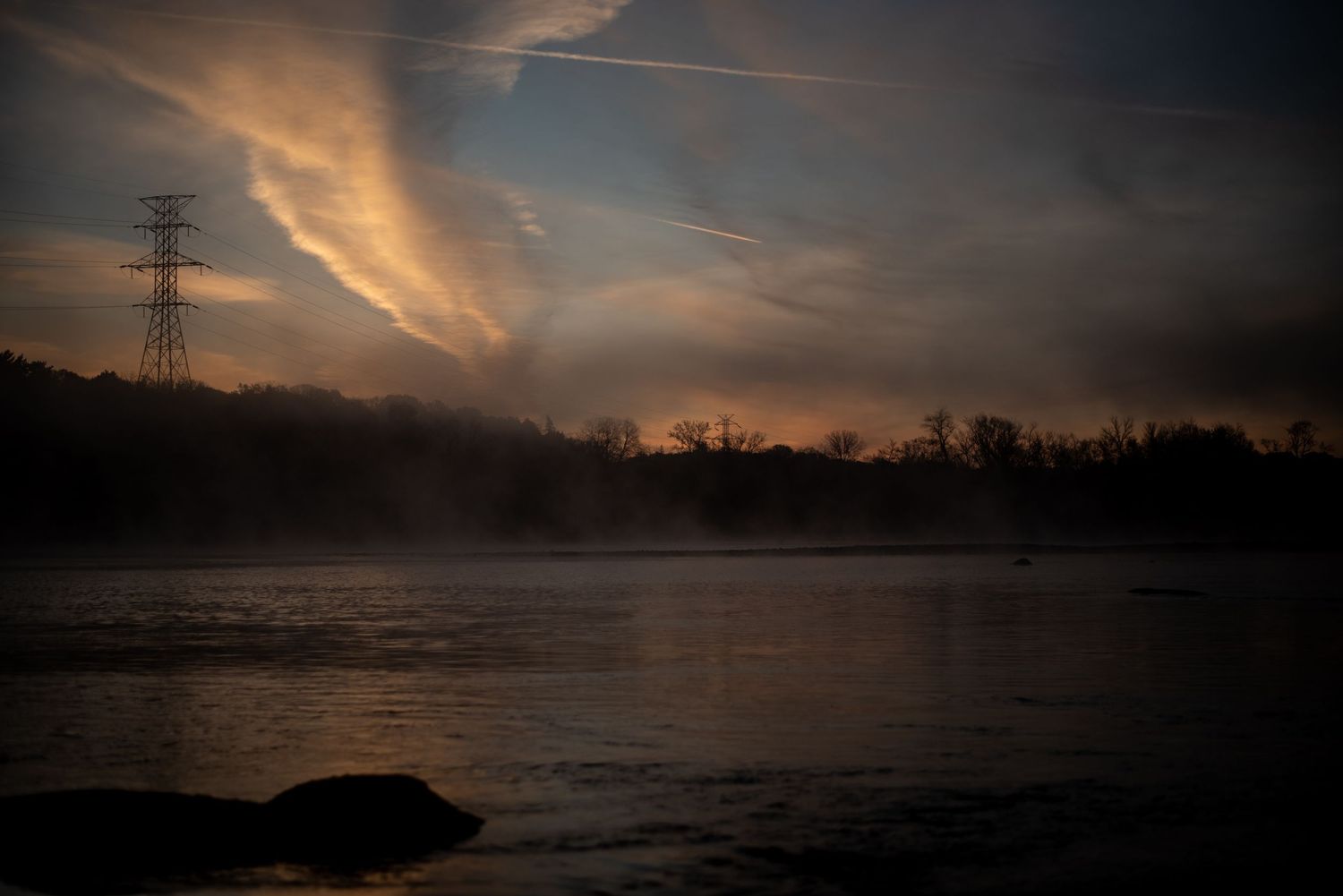 Dramatic sunset sky over a misty lake with silhouetted power lines and trees in the background.