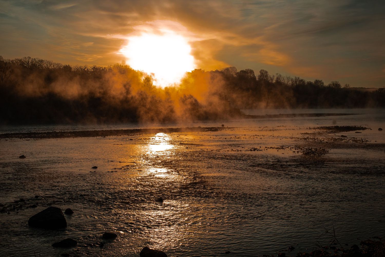 Dramatic winter sunrise over misty river with steam rising from water as golden sun reflects on icy surface.