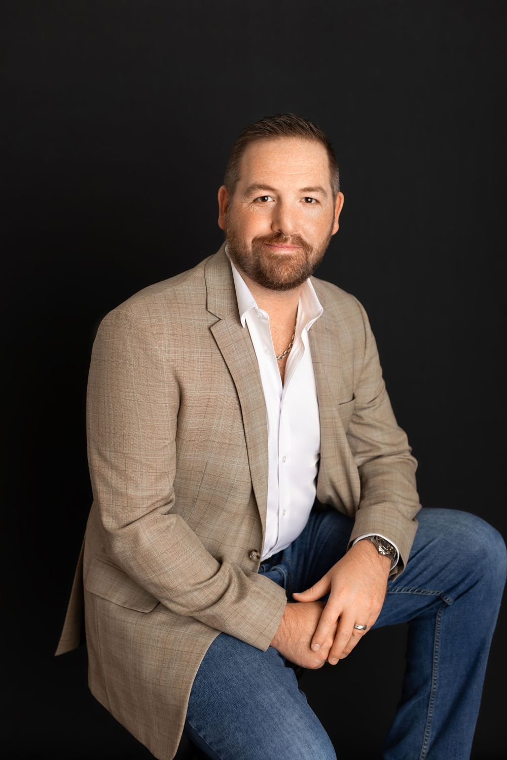 Professional portrait of person in beige blazer and white shirt seated against dark background.