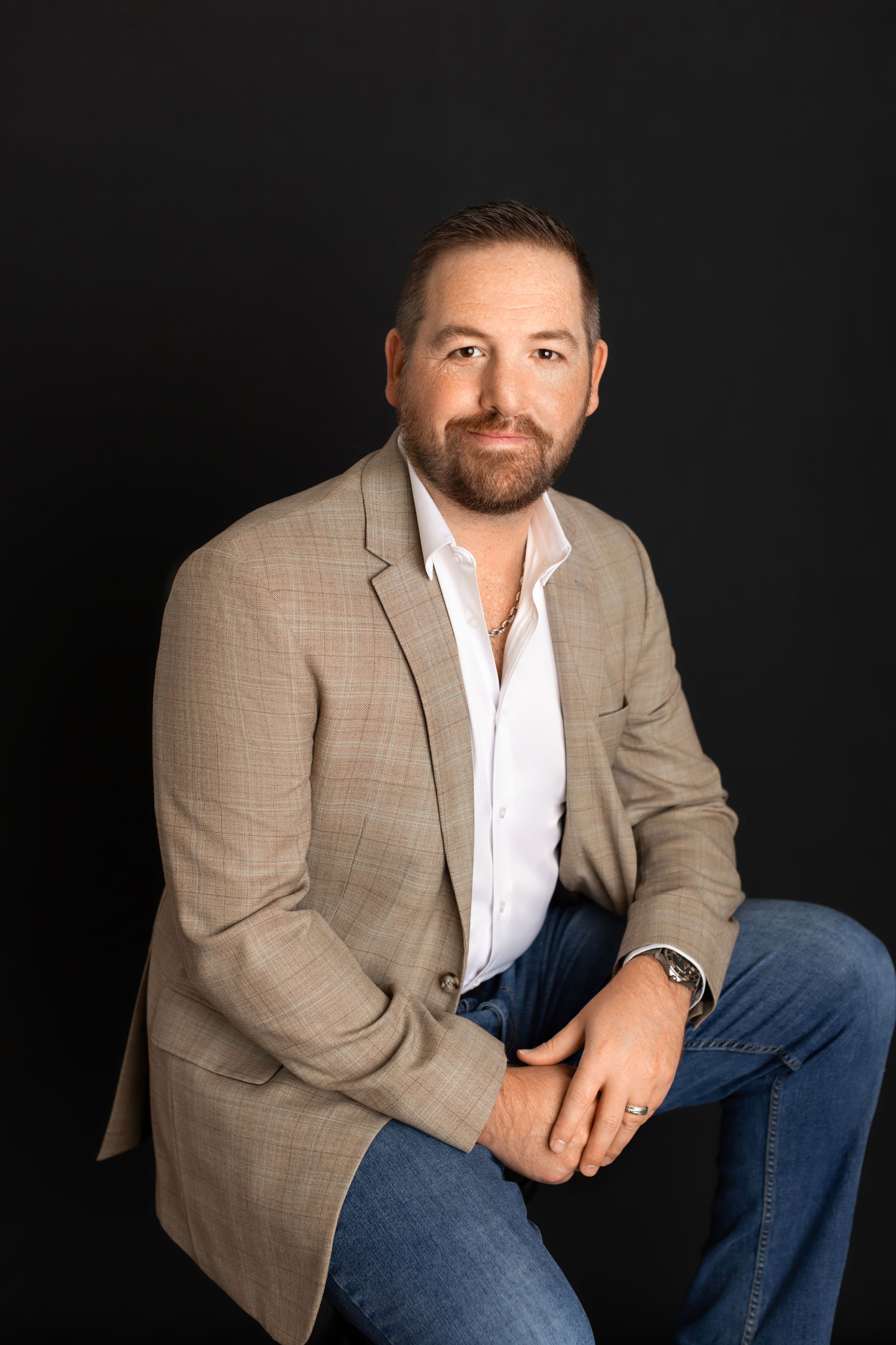 Professional portrait of person in beige blazer and white shirt seated against dark background.