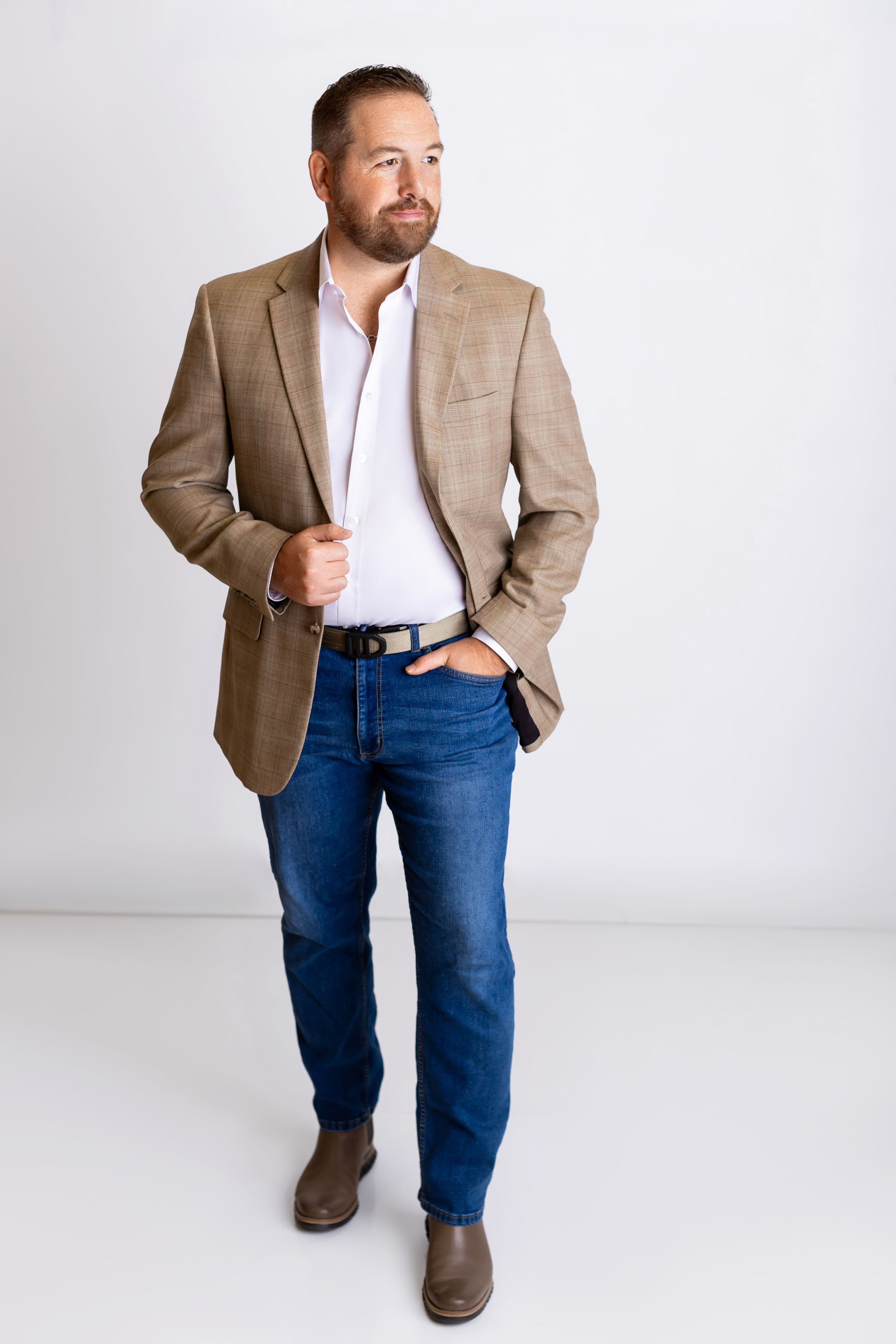 Smart casual outfit features beige blazer with white shirt, blue jeans and brown shoes on light background.