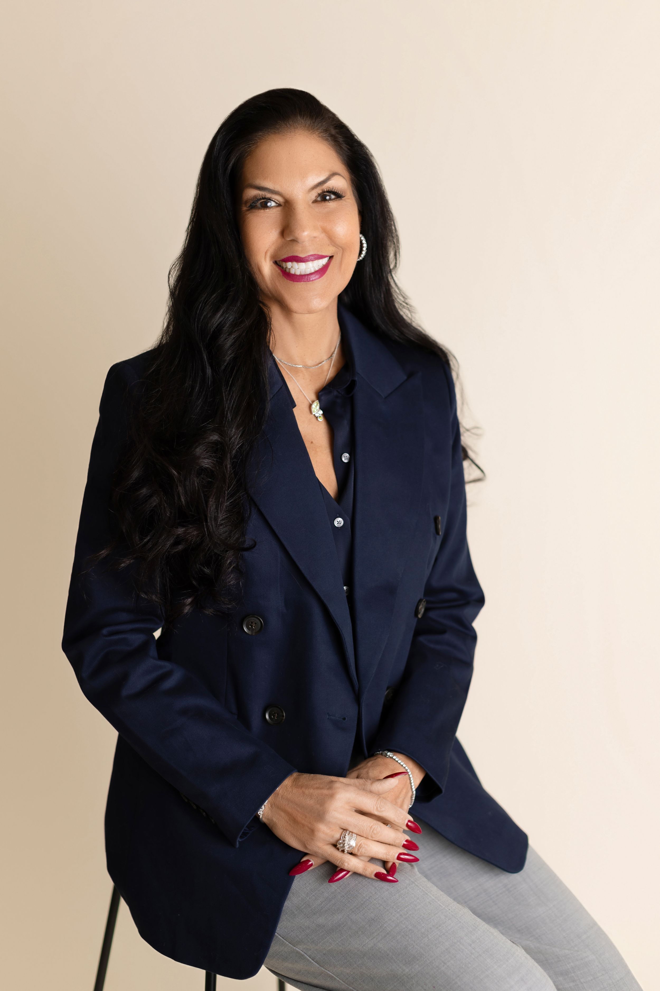 Professional portrait of someone wearing a navy blazer and grey pants while sitting and smiling warmly against beige background.