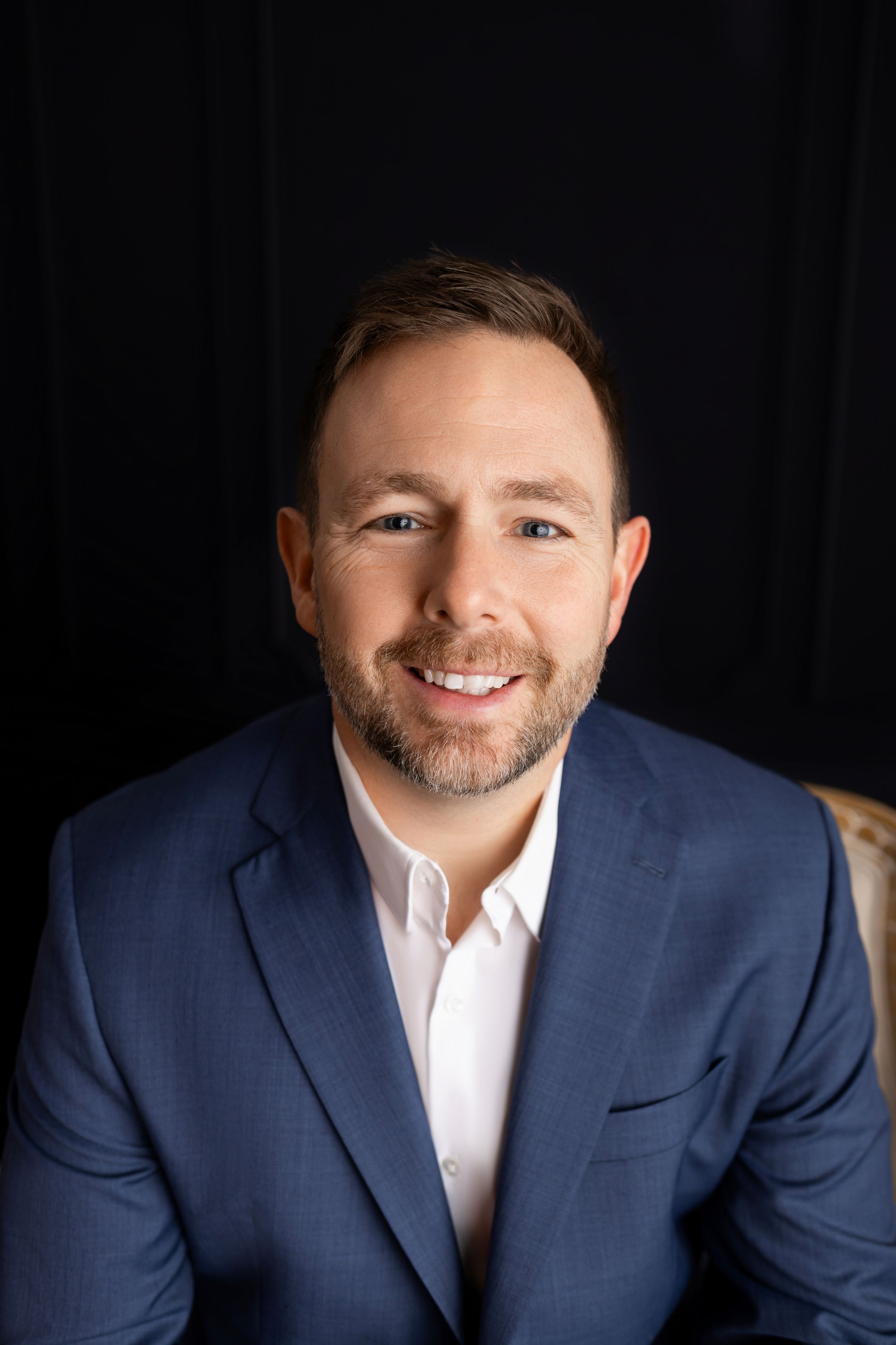 Professional headshot of someone in a navy blue suit jacket and white dress shirt against a dark background.