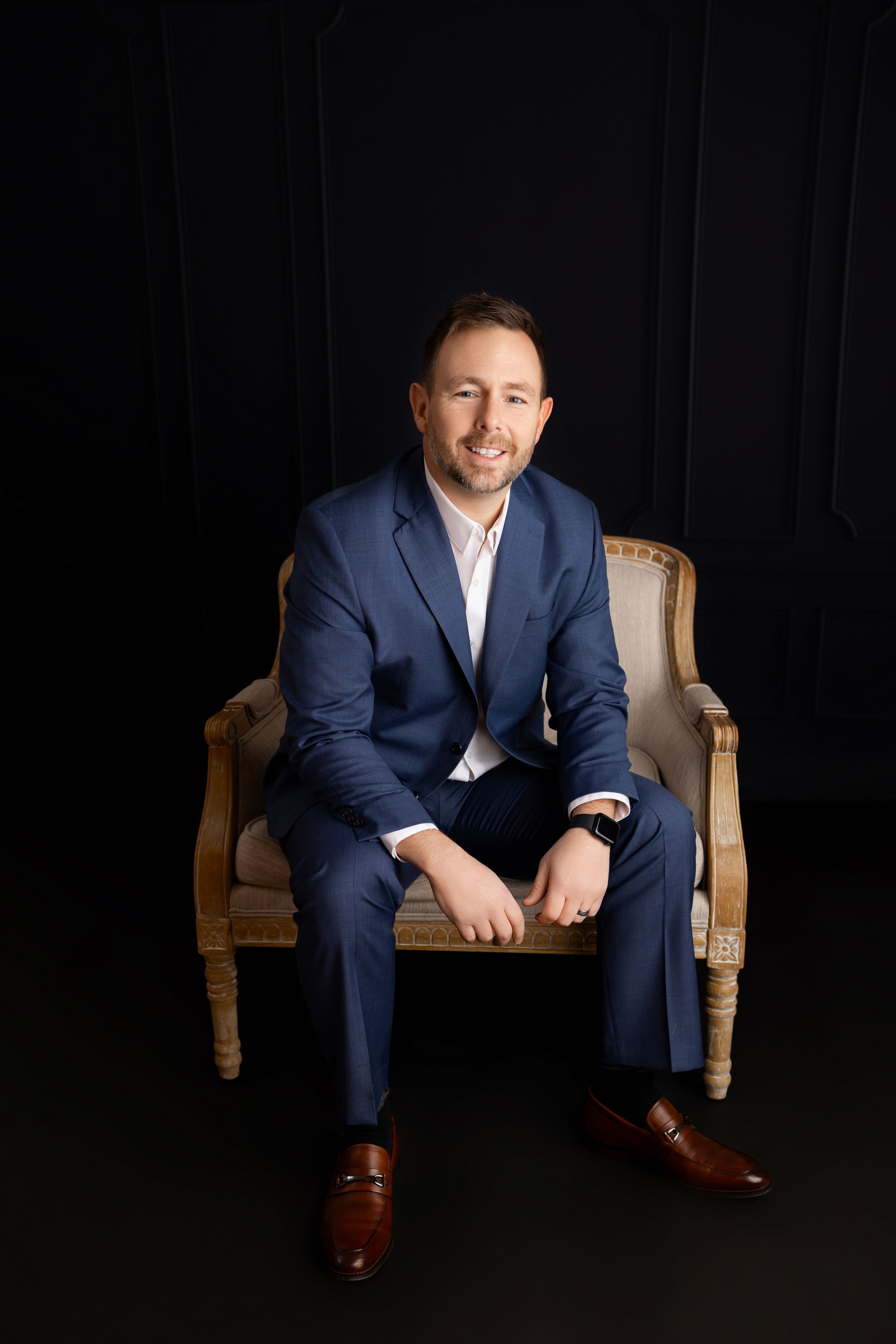 Professional business portrait of someone in a navy suit seated on an antique chair against a black backdrop.