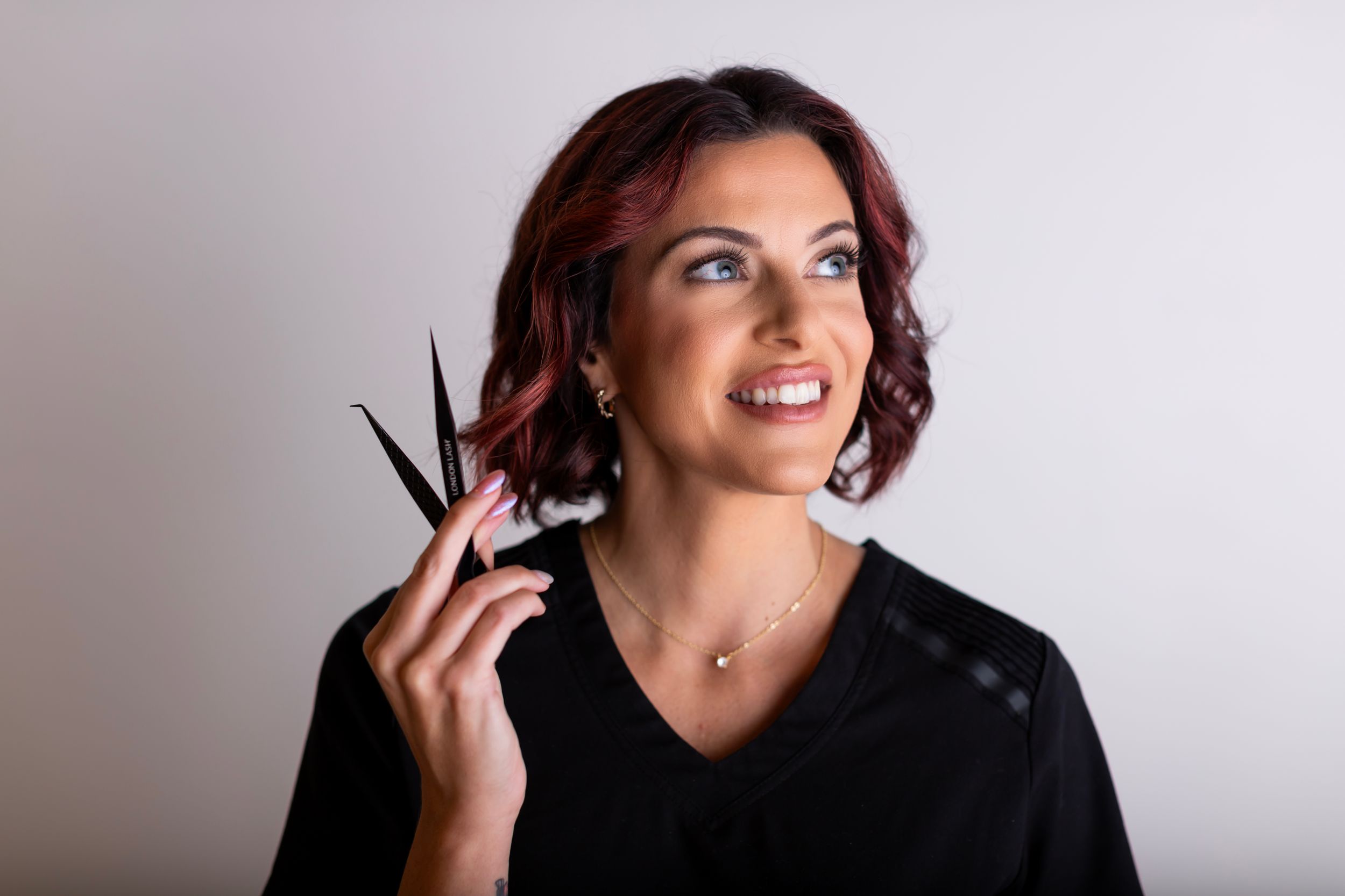 Professional hairstylist in black holding sharp scissors while looking up with a bright smile.