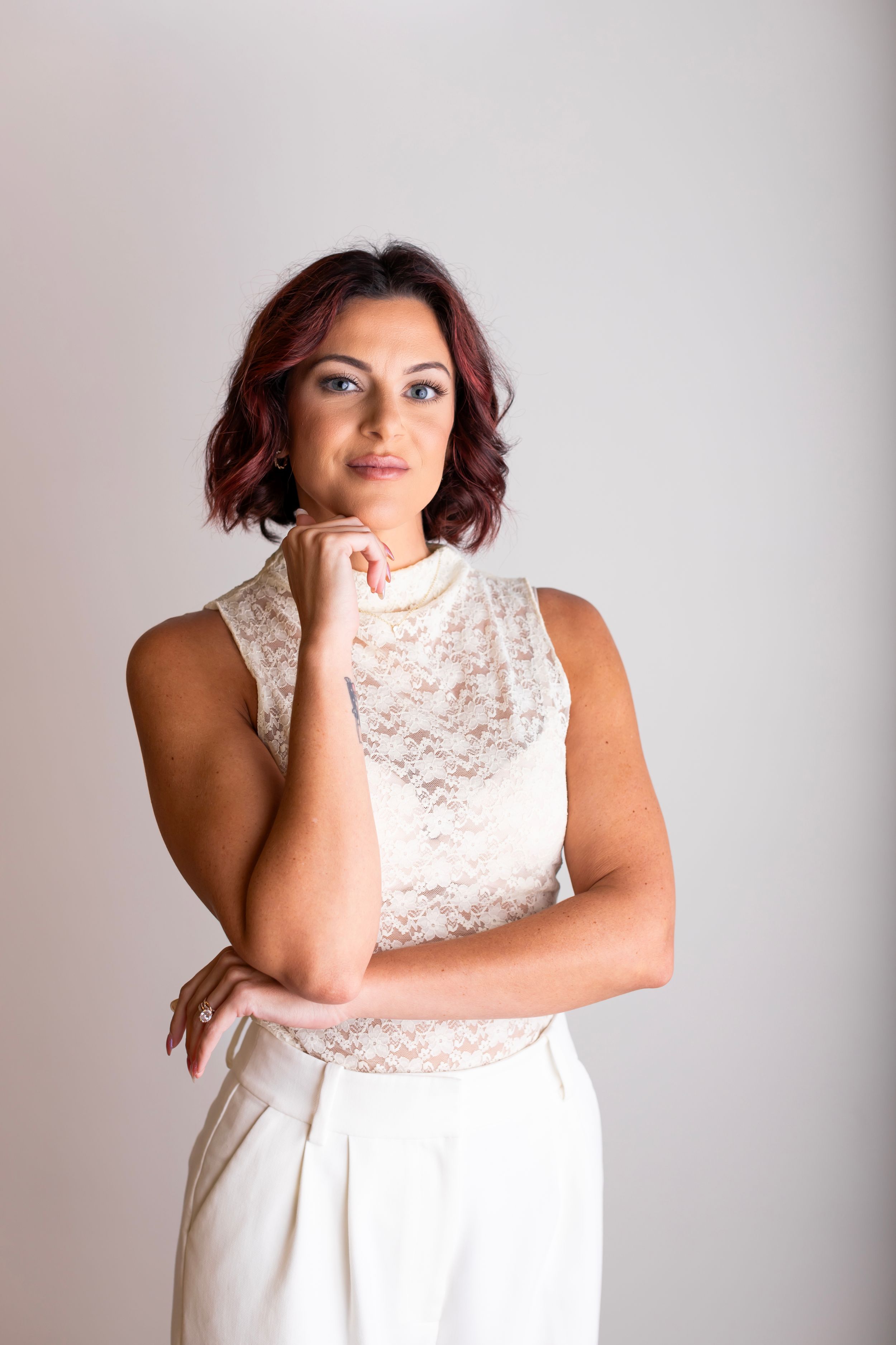 Person in sleeveless white lace dress poses with hand under chin against light background.