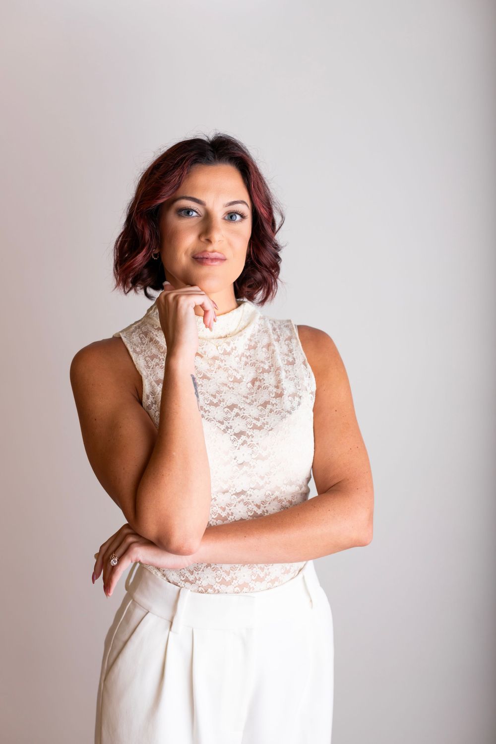 Person in sleeveless white lace dress poses with hand under chin against light background.