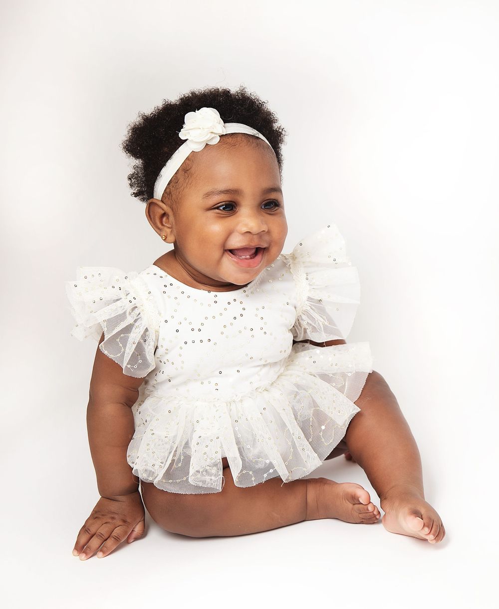 Smiling baby girl in white sparkly tutu dress and floral headband sitting against white background. During a baby photoshoot in Bexley