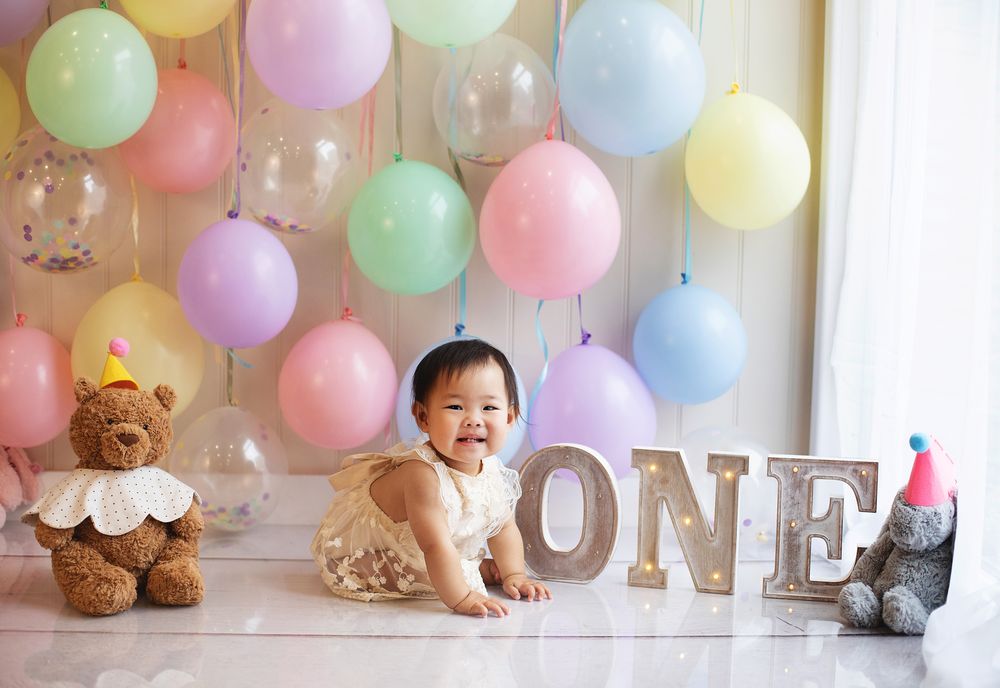 Baby girl smiles During a cake smash photoshoot in Bexley. Pastel balloons, teddy bear, and illuminated ONE sign.