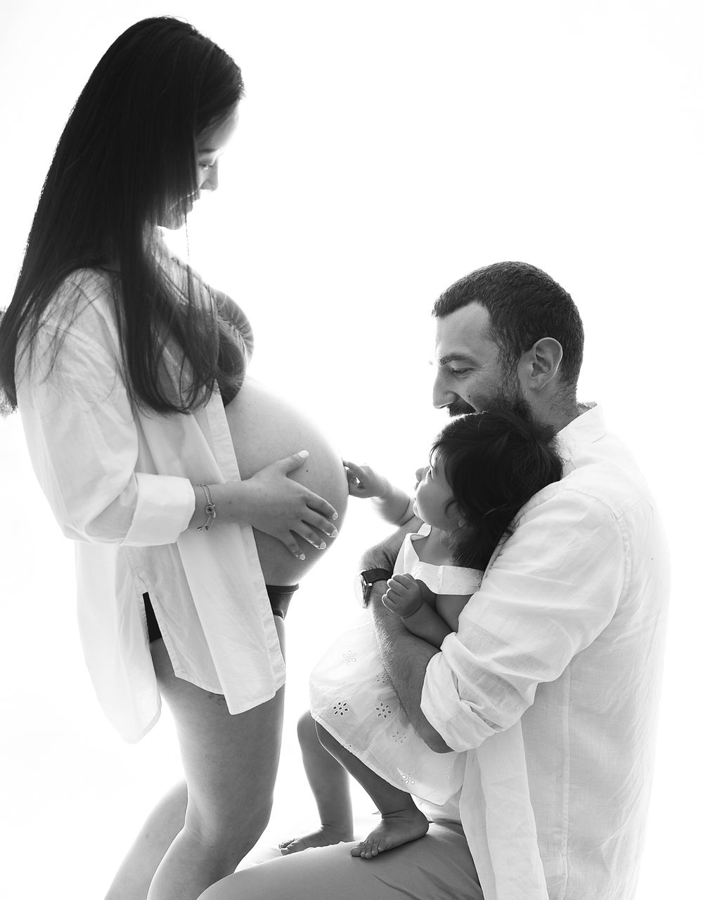 Black and white maternity photo of pregnant woman with partner and child touching baby bump in a maternity studio photoshoot in Bexley