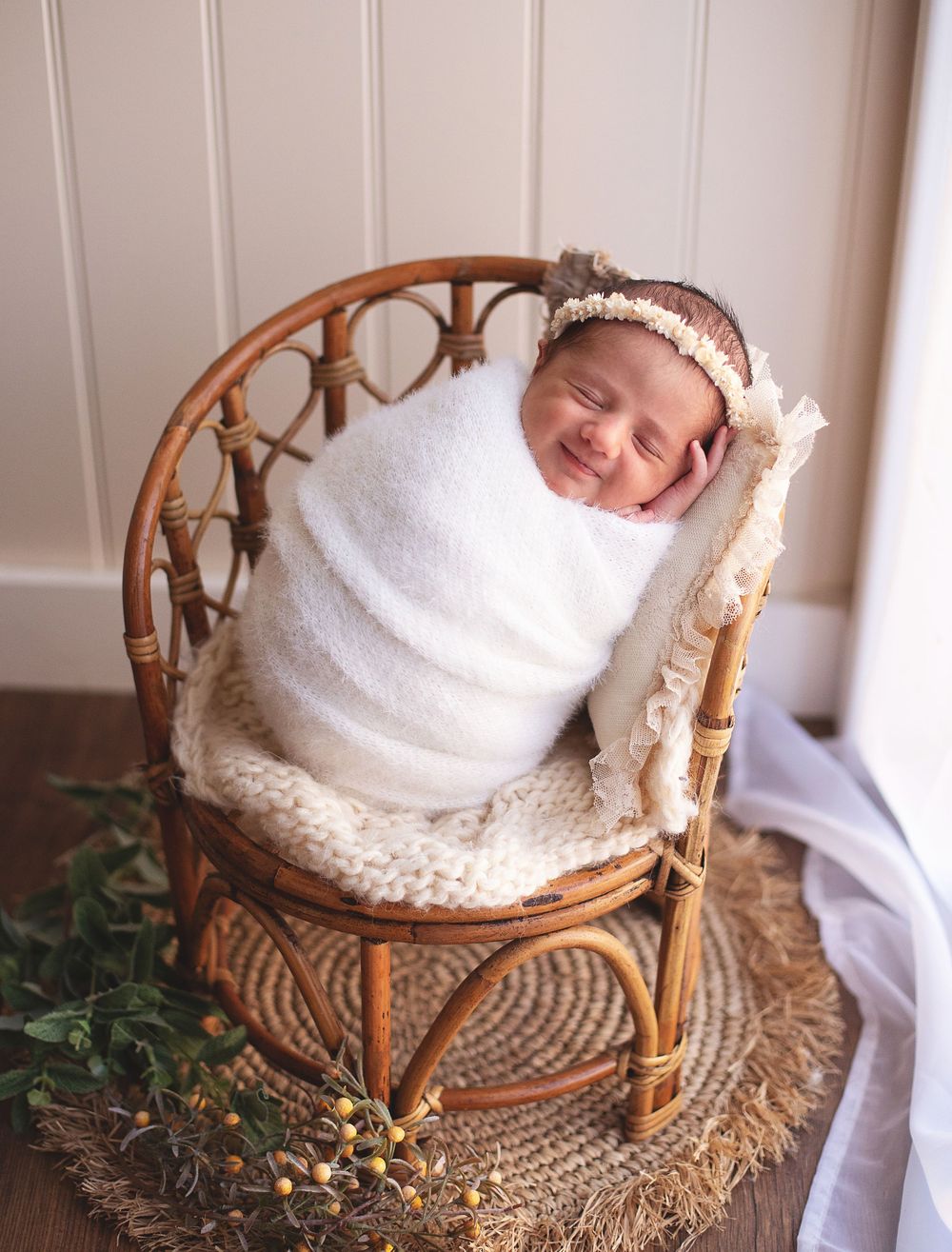 Smiling newborn baby swaddled in white wrap, wearing floral headband, posed in small rattan chair. During a newborn studio session in Bexley