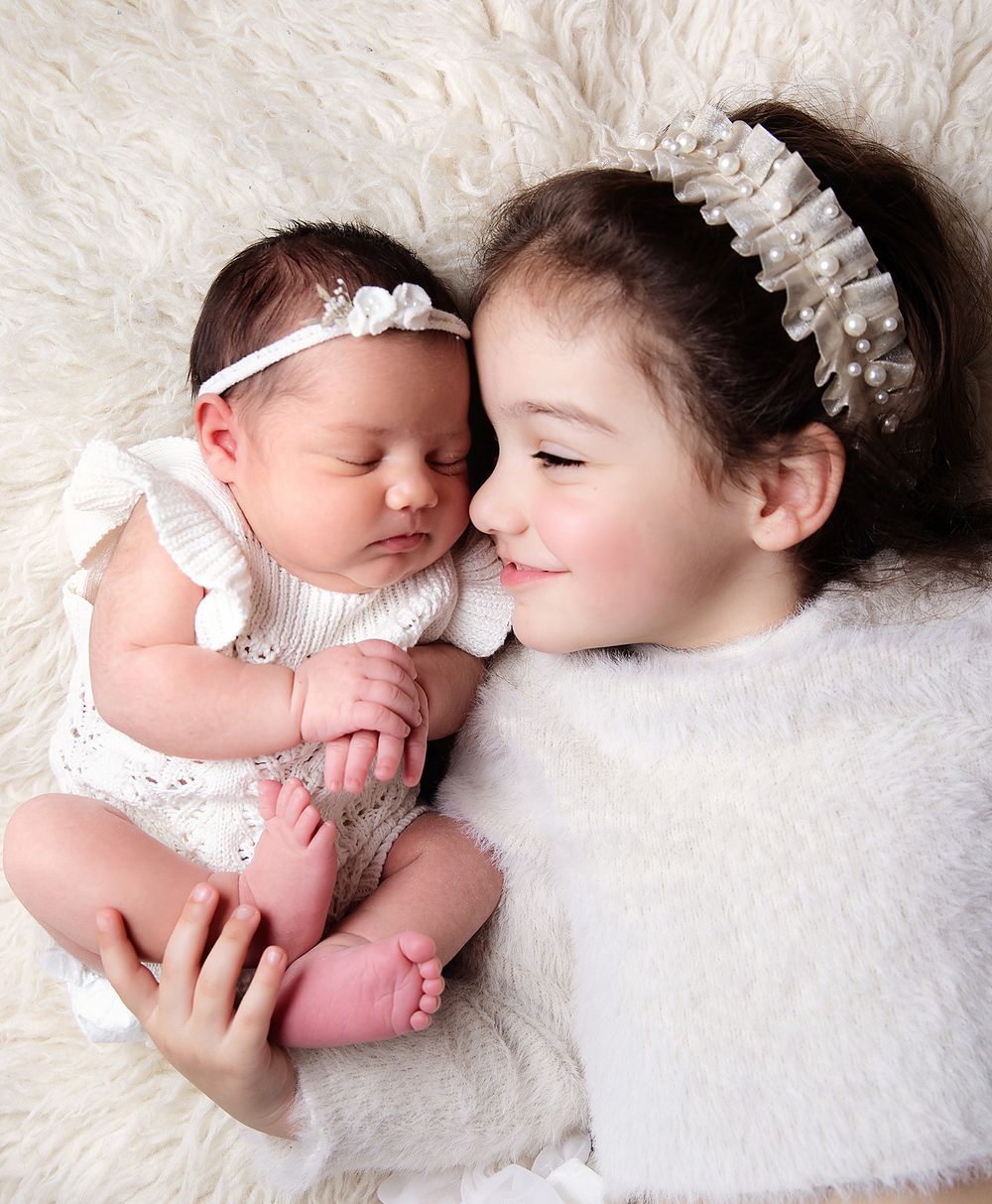 Newborn baby girl in white romper lying next to her older sister on a fluffy white blanket. During a newborn studio session in Bexley