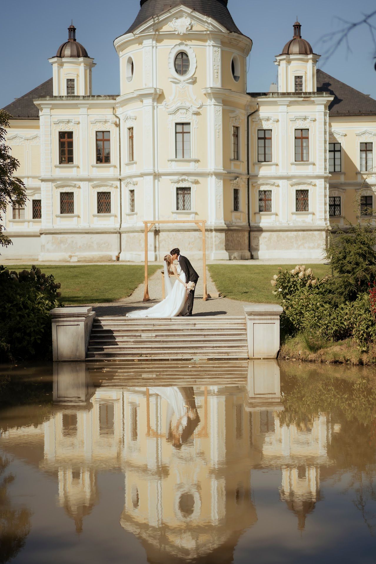 Elegant baroque castle with symmetrical towers and a reflective pond creating a perfect mirror image of a romantic wedding scene.