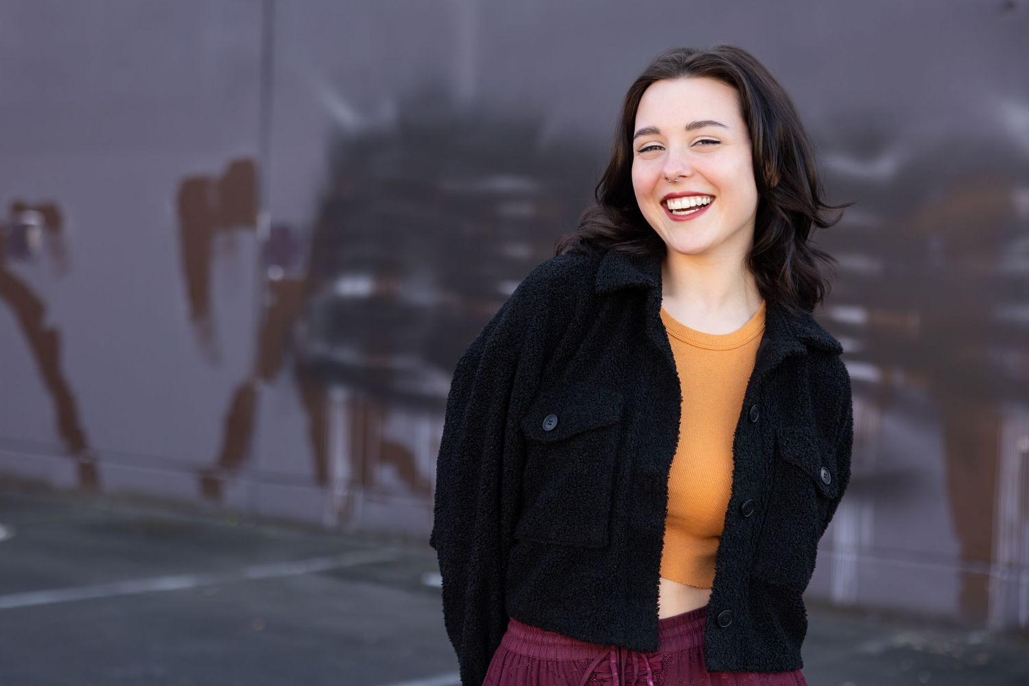 Person wearing black jacket and orange top laughs joyfully while standing outdoors against blurred urban background.