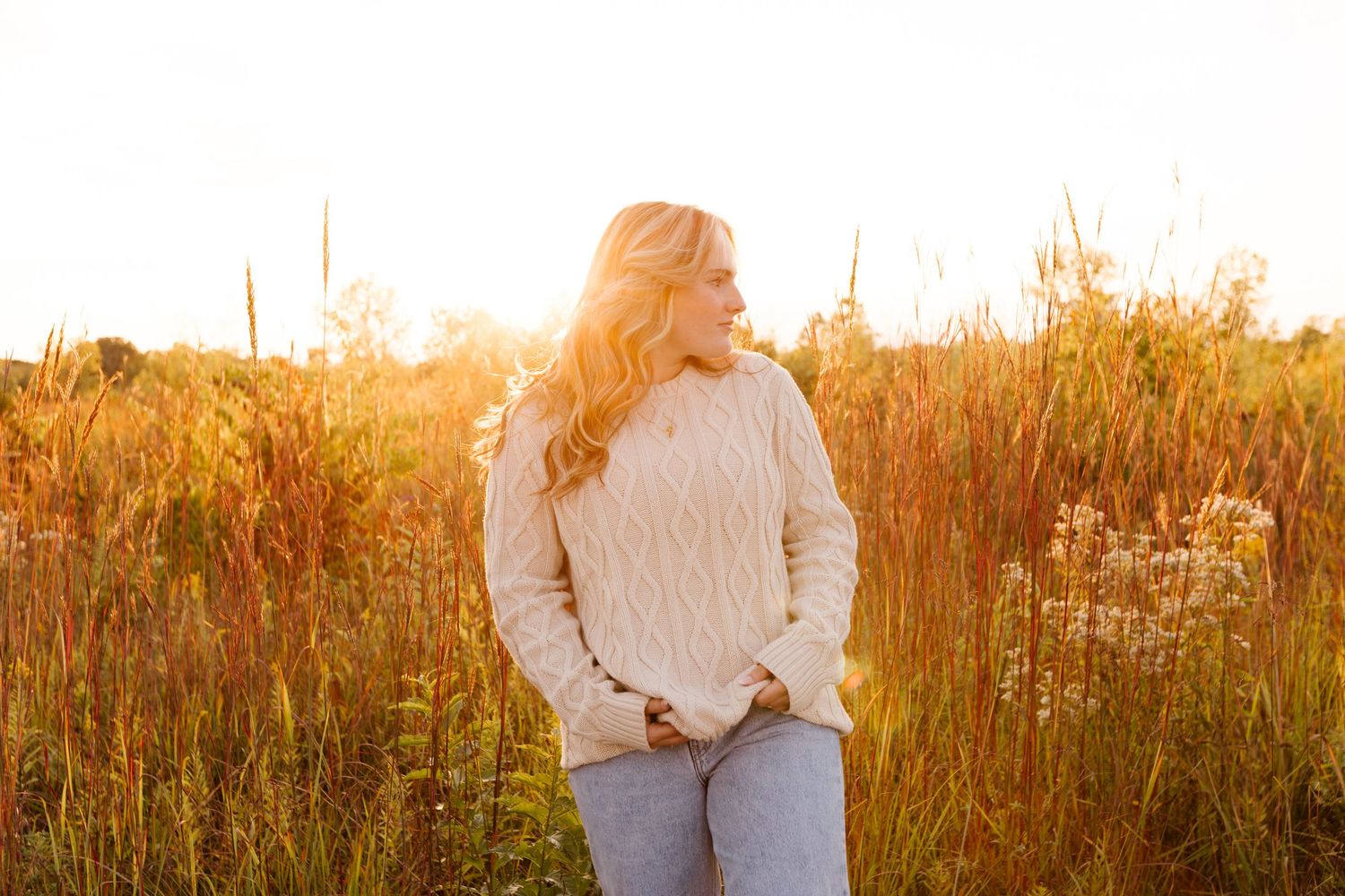 Person in white sweater standing in golden sunlit field of tall grass during autumn sunset.