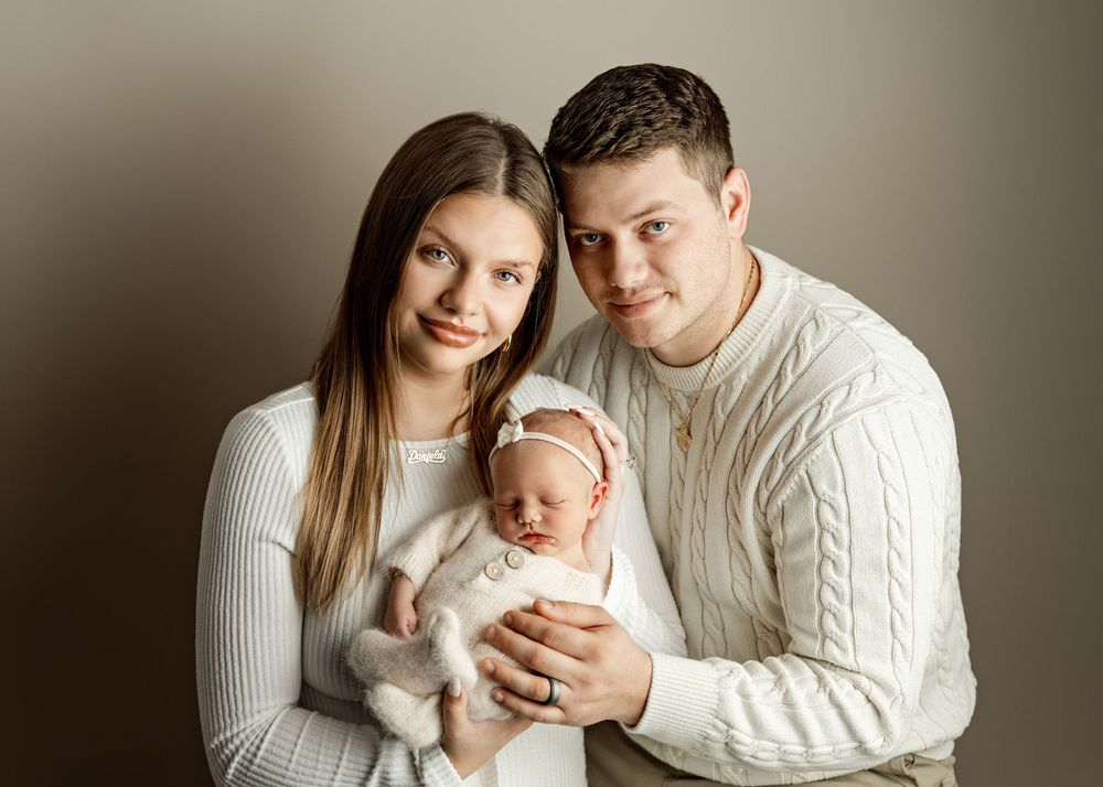 A couple in cream sweaters smiles warmly while holding their newborn baby during a professional studio portrait session.