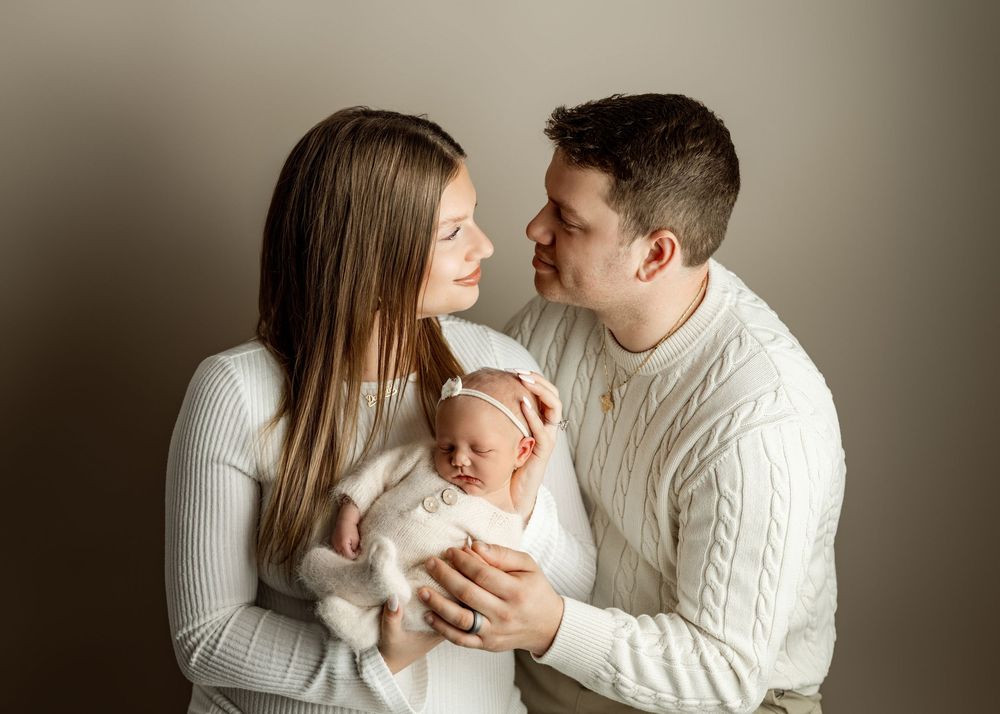 A couple in matching cream sweaters tenderly holds their newborn baby during a professional studio portrait session.