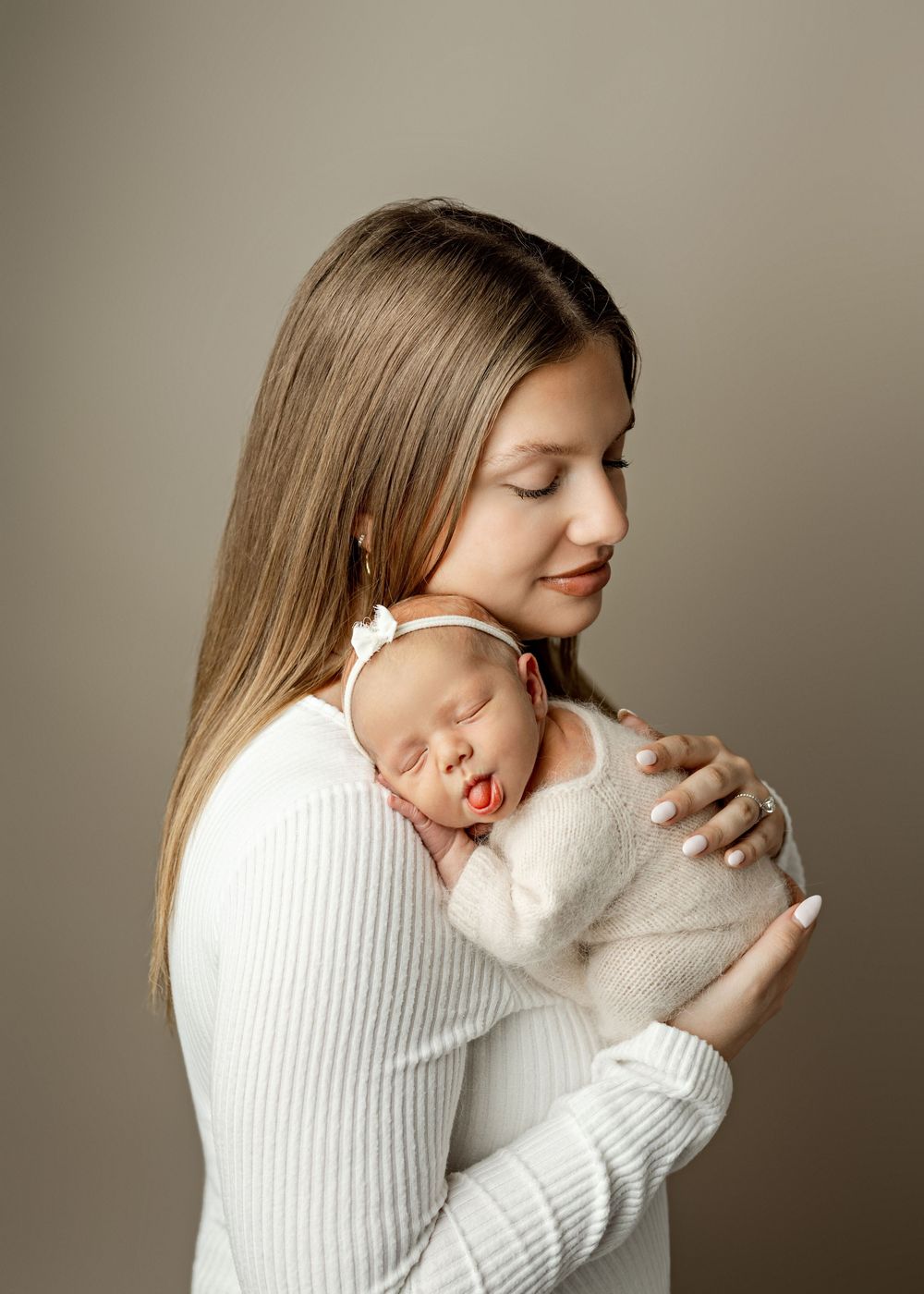 A mother in white gently cradles her sleeping newborn baby against her shoulder in a soft studio portrait.