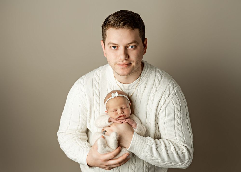 Man in white cable-knit sweater tenderly holds sleeping newborn baby against neutral studio background.