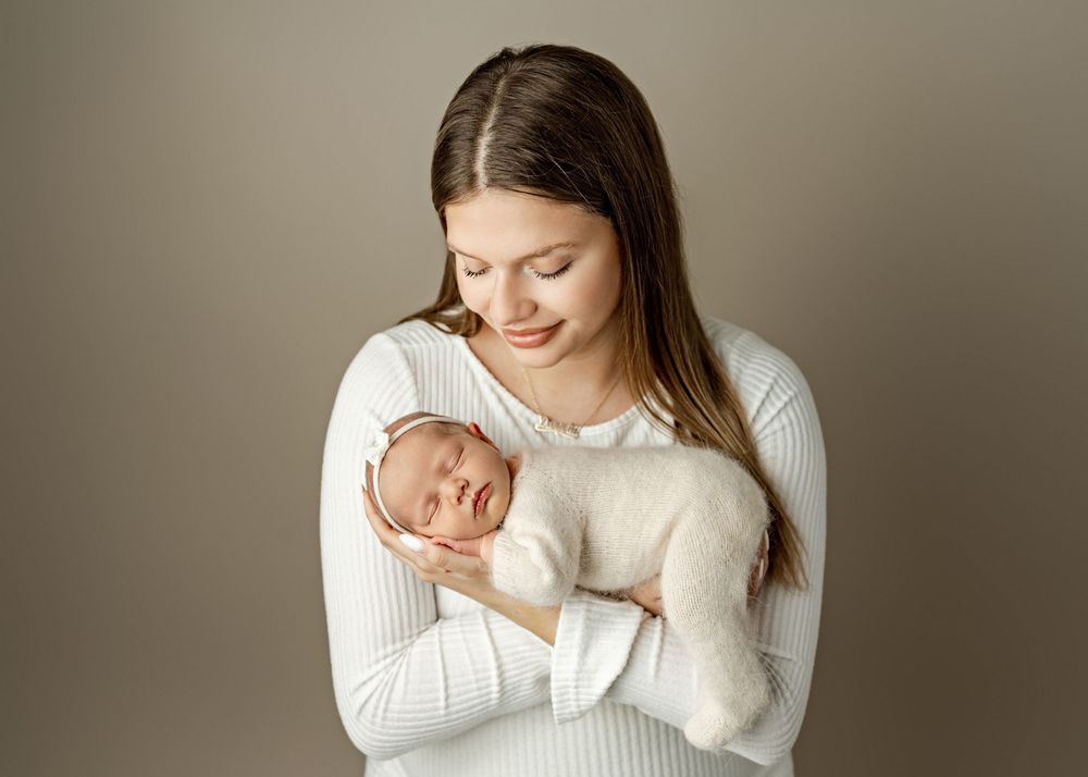 A young mother in a white dress tenderly holds her sleeping newborn baby wrapped in a cream swaddle.