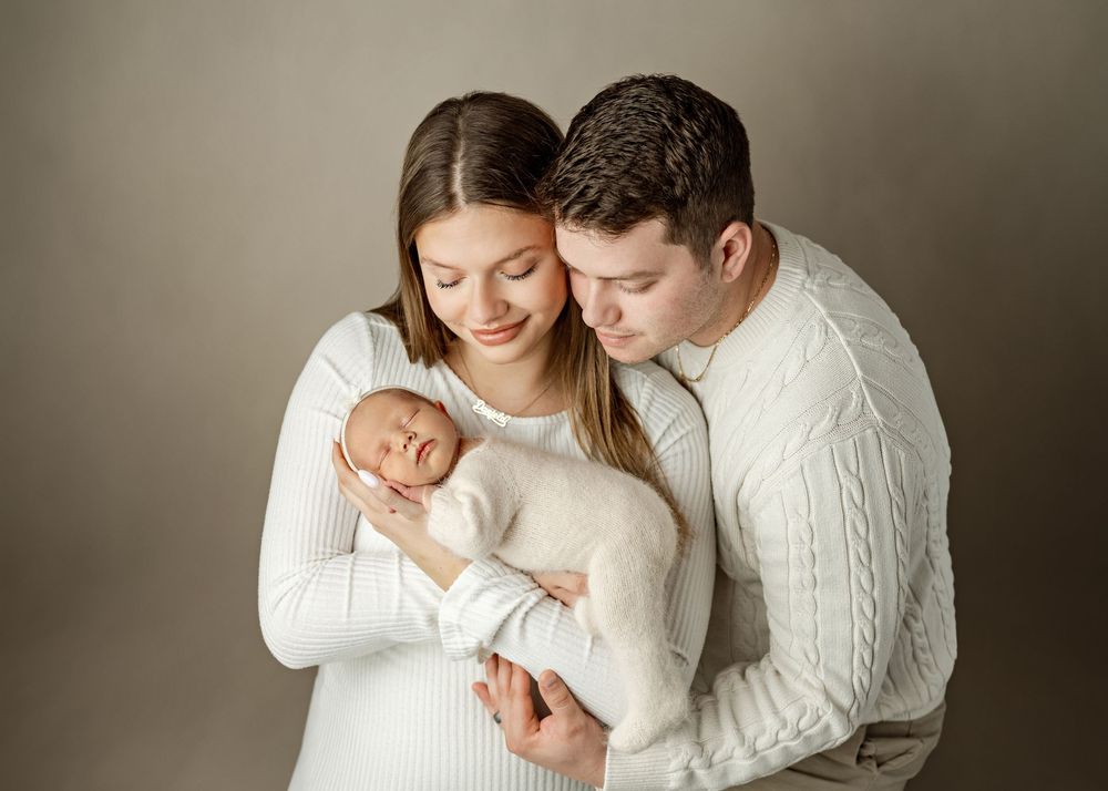New parents in white outfits tenderly hold their sleeping newborn baby wrapped in a soft white swaddle.
