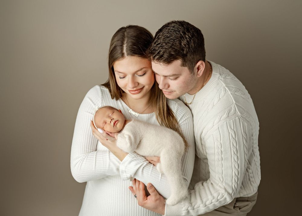 Parents in white outfits lovingly cradle their sleeping newborn baby in a professional studio portrait.