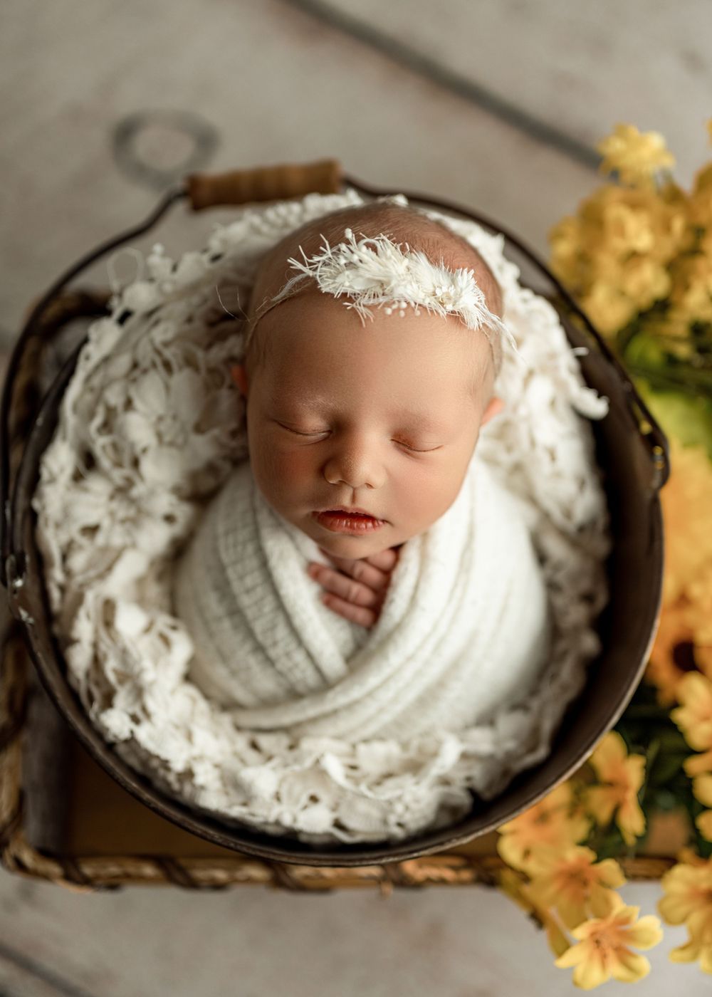Newborn baby swaddled in white wrap with feather headband, sleeping peacefully in a round metal bucket with yellow flowers.