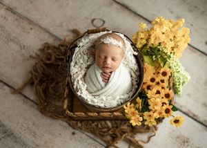 Newborn baby wrapped in white swaddle sleeping in a rustic basket surrounded by yellow flowers.