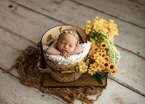 Newborn baby sleeping peacefully in a rustic basket surrounded by yellow flowers on a white wood floor.