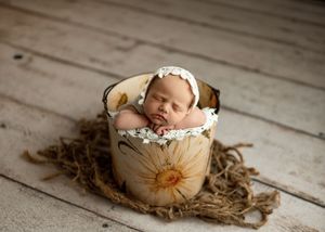 Sleeping newborn baby in a floral sunflower bucket with lace bonnet on rustic white wood floor.