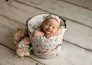 Sleeping newborn baby girl in floral bucket with pink flowers, wearing flower headband, newborn photography prop.
