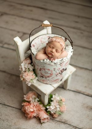 Sleeping newborn baby in floral bucket on white chair with pink flowers, professional newborn photography prop setup.