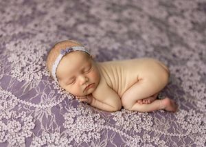 Sleeping newborn baby in beige wrap on purple lace blanket wearing a delicate floral headband.