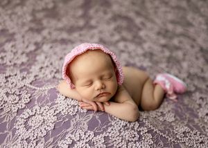 Sleeping newborn baby wearing a pink knit bonnet and booties on a purple lace floral backdrop.