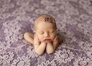 Sleeping newborn baby posed with hands under chin on purple lace blanket wearing a delicate floral headband.