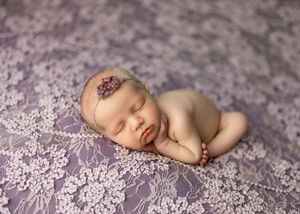 Newborn baby sleeping peacefully on purple lace fabric wearing a delicate floral headband in a professional photo.