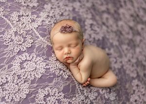 Newborn baby sleeping peacefully on purple lace fabric wearing a delicate floral headband in a professional photo.